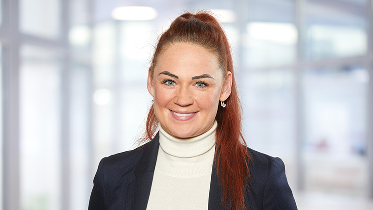 Eine Dame mit roten Haaren mit einem weißen Rollkragenpullover und einem schwarzen Sakko schaut lächelt in die Kamera